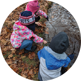 Three children gather around the pond and use sticks to interact with the pond’s surface