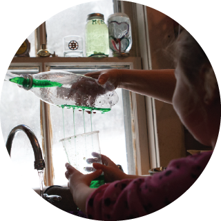 A girl experiments with a bottle of green liquid and a cup above the sink